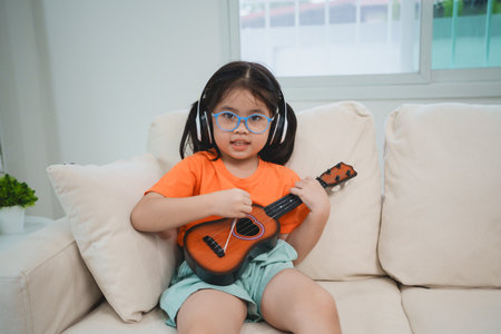 Asian baby girl child wearing glasses and headphone learning small guitar online study online on sofa in living room at home. Education learning music from home concept. Music for child concept.の写真素材
