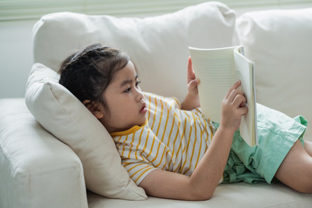 Joyful happy asian child baby girl smiling and  reading book while sitting on couch sofa in living room at home. Girl relex reading book smile at sofa In the house. Back to school concept.の写真素材