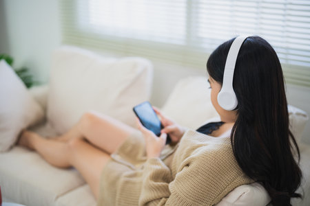 Asian freelance woman smile lying relax and using mobile and wearing headphone listen to music on sofa couch. Entrepreneur woman working searh job or trade stock or browse internet living room home.の写真素材