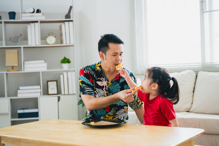 Asian happy children girl and her father feeding eating pizza and smiling in the living room at home. Children girl and her dad eating and tasting italian homemade pizza in house.の写真素材