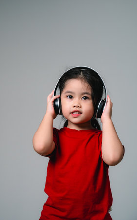 Cheerful happy asian children girl with black hair listening to music songs in headphones on a gray background. Portrait cute girl waring red shirt and looking at camera studio lighting.の写真素材