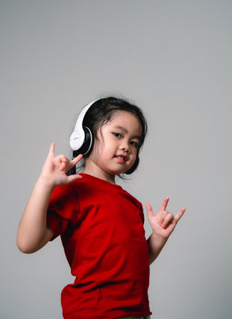 Cheerful happy asian children girl with black hair listening to music songs in headphones on a gray background. Portrait cute girl waring red shirt and looking at camera studio lighting.の写真素材