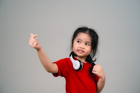 Cheerful happy asian children girl with black hair listening to music songs in headphones on a gray background. Portrait cute girl waring red shirt and looking at camera studio lighting.の写真素材