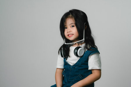 Portrait of relaxed cute little asian girl child funny smiling and laughing wearing white headphones and glasses, keeps hands on her ears, posing and smiling enjoying isolated over gray background.の写真素材