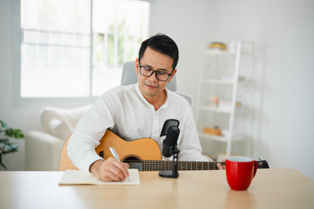 Asian man wearing headphone play guitar and sing a song and learning online with laptop and notesbook and compose writing song, record sound and use computer recording music program. Musician concept.の写真素材