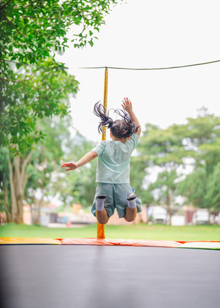 Asian child girl is jumping on trampoline on playground background. Happy laughing kid outdoors in...