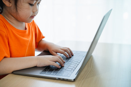 Close up hand. Asian baby girl wearing orange t-shirt use laptop and study e-learning online on wood table desk in living room at home. Education learning online from home concept.の写真素材