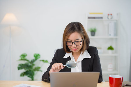 Asian businesswoman wearing suit and glasses holding magnifying glass and working laptop. Seaching...
