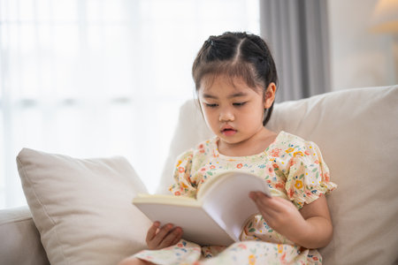 Joyful happy asian child baby girl smiling and reading book while sitting on couch sofa in living room at home. Girl relex reading book smile at sofa In the house. Back to school concept.の写真素材