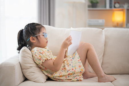 Joyful happy asian child baby girl smiling and reading book while sitting on couch sofa in living...