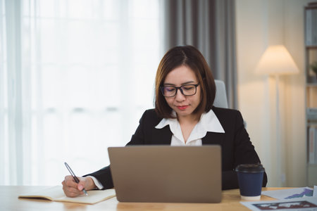 Asian businesswoman wearing suit and writing notes book and working laptop on table at home. Entrepreneur woman working for business at living room home. Business work home concept.の写真素材