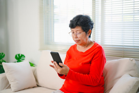 Happy senior old asian woman enjoying using mobile apps texting typing messages sit on sofa, smiling old lady holding smartphone looking smartphone browsing social media or learning technology at homeの写真素材
