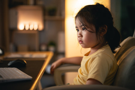 Asian baby girl wearing t-shirt concentrate to use laptop and study online on wood table desk in living room at home at night. Education learning online from home concept.の写真素材