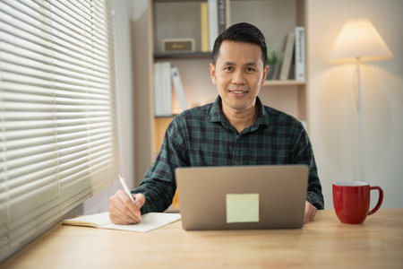 Asian freelance man happy relax and smile working typing on laptop and write notes in notebook at...