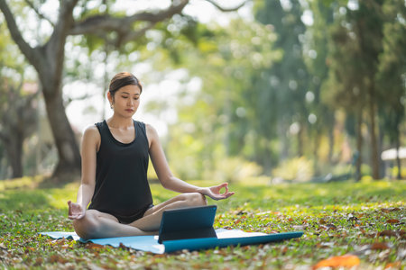 Young asian woman learning or teaching yoga exercise on a video conference outdoor sitting on yoga mat in the park, sport yoga concept.の写真素材