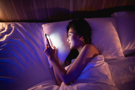 Asian woman using mobile phone smartphone laying on the bed in the bedroom. Women scrolling social networks on mobile. Content female lying in bed under blanket and watching movie on smartphone.の写真素材