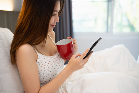 Joyful asian woman drink hot coffee and using mobile phone while sitting on the bed in bedroom at home. Asian women relex laying smile after wake up on the bed house. Activity hobby at house concept.の写真素材