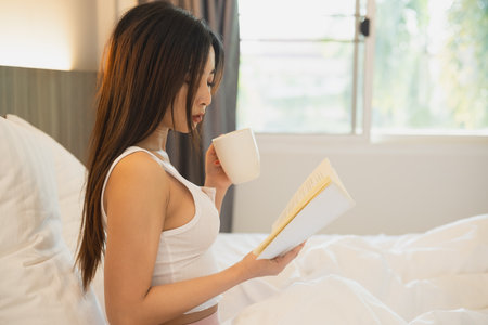 Joyful asian woman drink hot coffee and reading a book while sitting on the bed in bedroom at home. Asian women relax laying smile after wake up on the bed house. Activity hobby at house concept.の写真素材