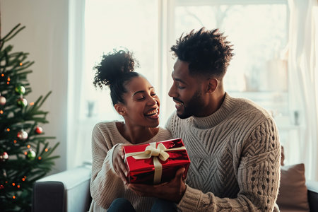 American african lover. Man presenting gift to his girlfriend in room at home. Symbol of their love. Man looks tenderly at his beloved. Valentine's day celebration.の素材