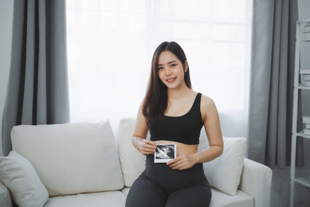 Pregnant woman keeping hand on belly and holding ultrasound image at living room home. Family mother mom pregnant concept.の写真素材