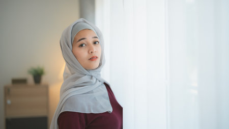 Portrait of beautiful asian muslim woman in a shoulder-length hijab standing side window in the living room with large windows. Oriental women smiling brightly posing in a modern home.の写真素材