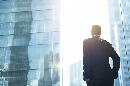 View from behind back. Male businessman standing looking at a large building with glass. Concept of success, work, vision, thinking, strategy, planning, achieving goals.の素材