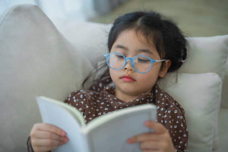 Happy asian child baby girl smiling wearing glasses and reading book while sitting on couch sofa in living room at home. Girl relex reading book smile at sofa In the house. Back to school concept.の写真素材