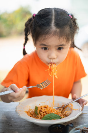 Young Girl children Enjoying a Forkful of Spaghetti. Close-up of a little girl in an orange shirt eating spaghetti with a focused expression, outdoors restaurant.の写真素材