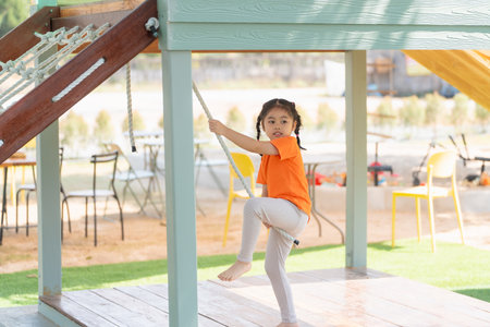 Young Girl Playing on a Rope Ladder. A little girl in an orange shirt climbs a rope ladder at a playground, looking back with concentration. Baby girl kids activity concept.の写真素材