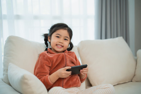 Happy Child Playing on Smartphone at Home. A joyful young girl with pigtails is engaged in playing a game on her smartphone while sitting comfortably on a sofa.の写真素材