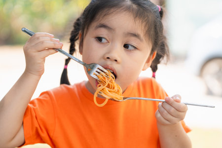Young Girl children Enjoying a Forkful of Spaghetti. Close-up of a little girl in an orange shirt eating spaghetti with a focused expression, outdoors restaurant.の写真素材