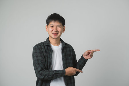 Confident Teen Man Boy with Braces Pointing Upwards. Smiling Asian teenager in checkered shirt pointing upwards on a neutral grey background. Portrait handsome man in studio concept.の写真素材