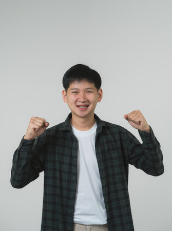 Cheerful Teen Boy with Braces Celebrating Victory. Joyful Asian teen boy with braces clenching fists in a triumphant gesture on a grey background. Portrait handsome man in studio concept.の写真素材