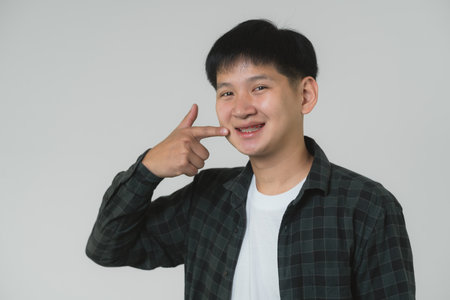 Happy Asian Teen Boy Pointing at Braces. Smiling Asian teenager man pointing at his dental braces on a light grey background. Portrait handsome man in studio concept.の写真素材