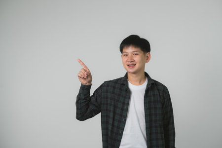 Confident Teen Man Boy with Braces Pointing Upwards. Smiling Asian teenager in checkered shirt pointing upwards on a neutral grey background. Portrait handsome man in studio concept.の写真素材