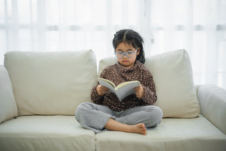Happy asian child baby girl smiling wearing glasses and reading book while sitting on couch sofa in living room at home. Girl relex reading book smile at sofa In the house. Back to school concept.の写真素材