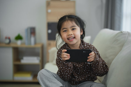 Asian child girl looking using and touch mobile phone screen on couch sofa. Baby smiling funny time to use mobile phone. Too much screen time. Cute girl watching videos while tv, Internet addiction.の写真素材
