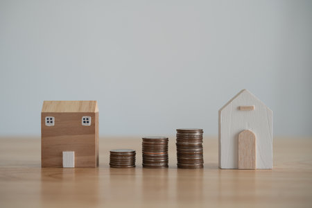 Real Estate Savings Concept with House Models and Coins. Two wooden house models beside rising stacks of coins on a clear surface, symbolizing investment growth. Finance home loan or interest concept.の写真素材