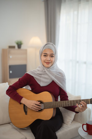 Muslim Islam woman wearing hijab or grey scarf is playing a guitar. She is smiling and she is enjoying herselfの写真素材