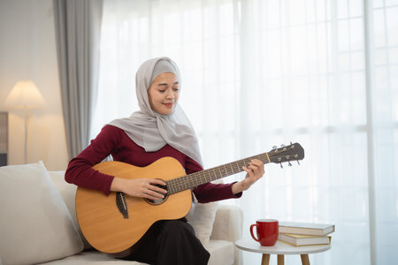 Muslim Islam woman wearing hijab or grey scarf is playing a guitar. She is smiling and she is enjoying herselfの写真素材