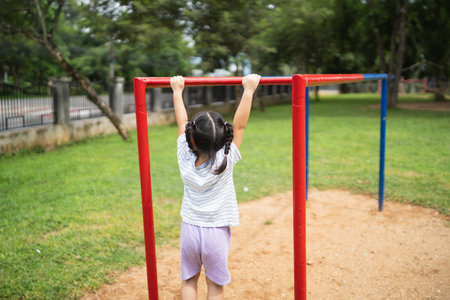 A little girl is hanging from a red and blue park bar. She is wearing a white shirt and purple...