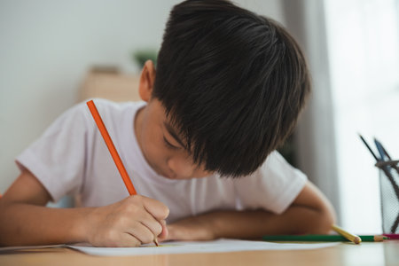 A young boy is sitting at a desk and writing with a pencil. He is focused on his work and he is in a serious mood. Concept of a child learning and practicing their writing skillsの写真素材