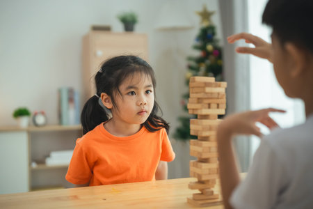 A girl in an orange shirt is sitting at a table with a stack of wooden blocks. A boy is standing behind her, watching her. Scene is playful and lightheartedの写真素材