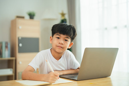A young boy sits at a table with a laptop and a pencil. He is focused on writing something in his notebookの写真素材