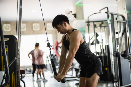A focused man in a black tank top and shorts performs a tricep pushdown exercise using a cable...