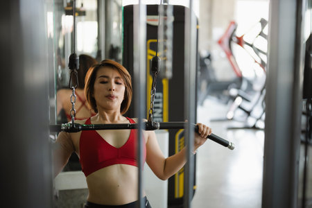 Sport determined woman in a red sports bra and black leggings uses a lat pulldown machine in a...