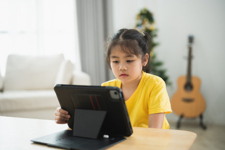Asian young girl is sitting at a table with a tablet in front of her. She is looking at the screen with a serious expression on her faceの写真素材