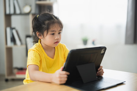 Asian young girl is sitting at a table with a tablet in front of her. She is looking at the screen with a serious expression on her faceの写真素材