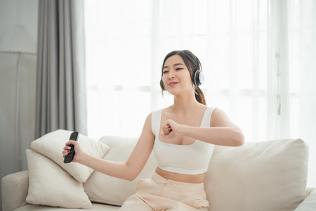Asian woman is sitting on a couch and playing a video game or dancing. She is wearing headphones and has a thumbs up gestureの写真素材