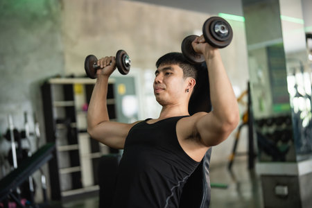 Asian man in a black tank top performs a seated dumbbell shoulder press in a modern gym. His muscular build and concentrated expression highlight his dedication to strength training.の写真素材